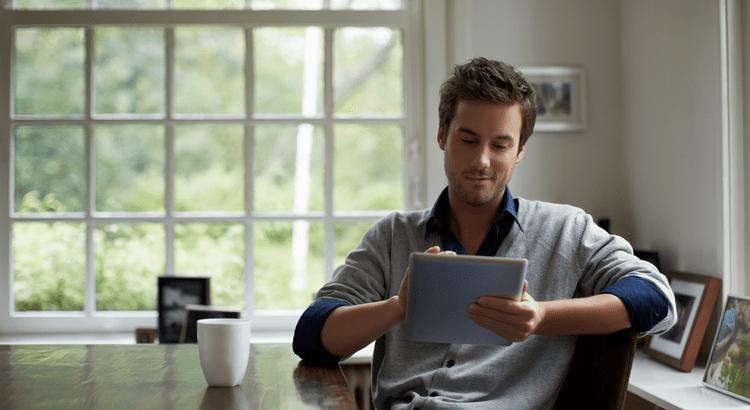 Person reading on a tablet at home near a large sunlit window with a coffee mug.